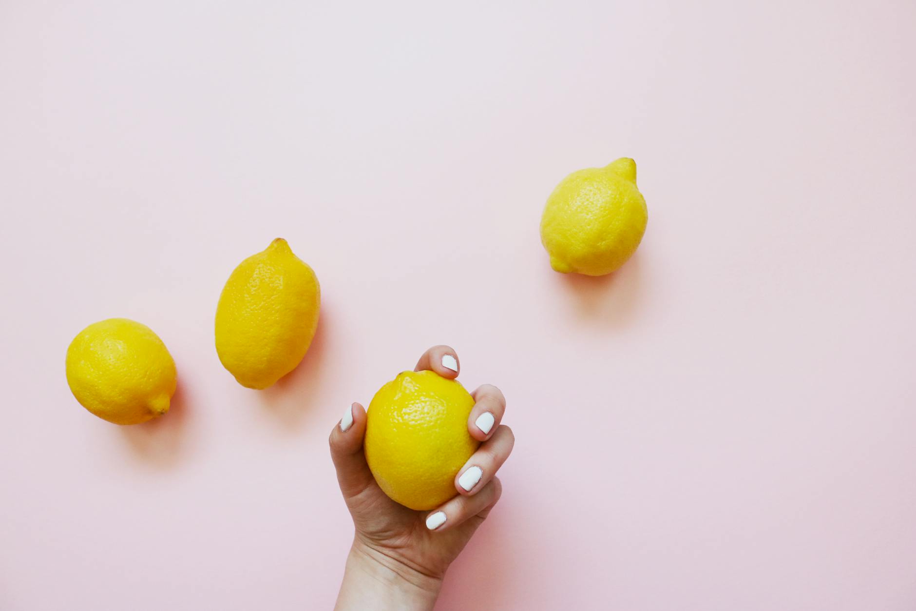 A hand with white nails holding a lemon on a soft pink background, surrounded by additional lemons.