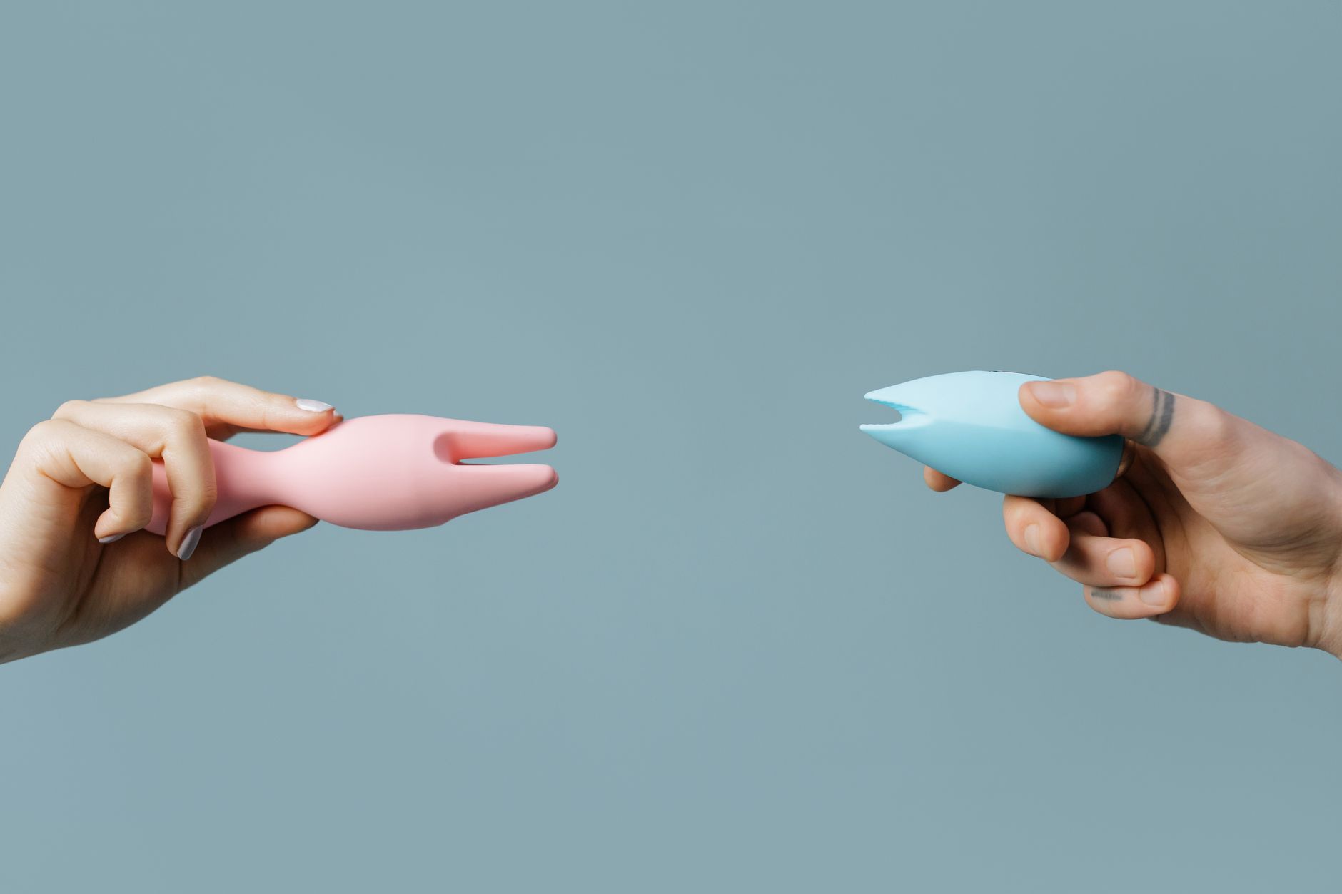 Two hands holding vibrators against a pastel background, illustrating clitoral pleasure devices
